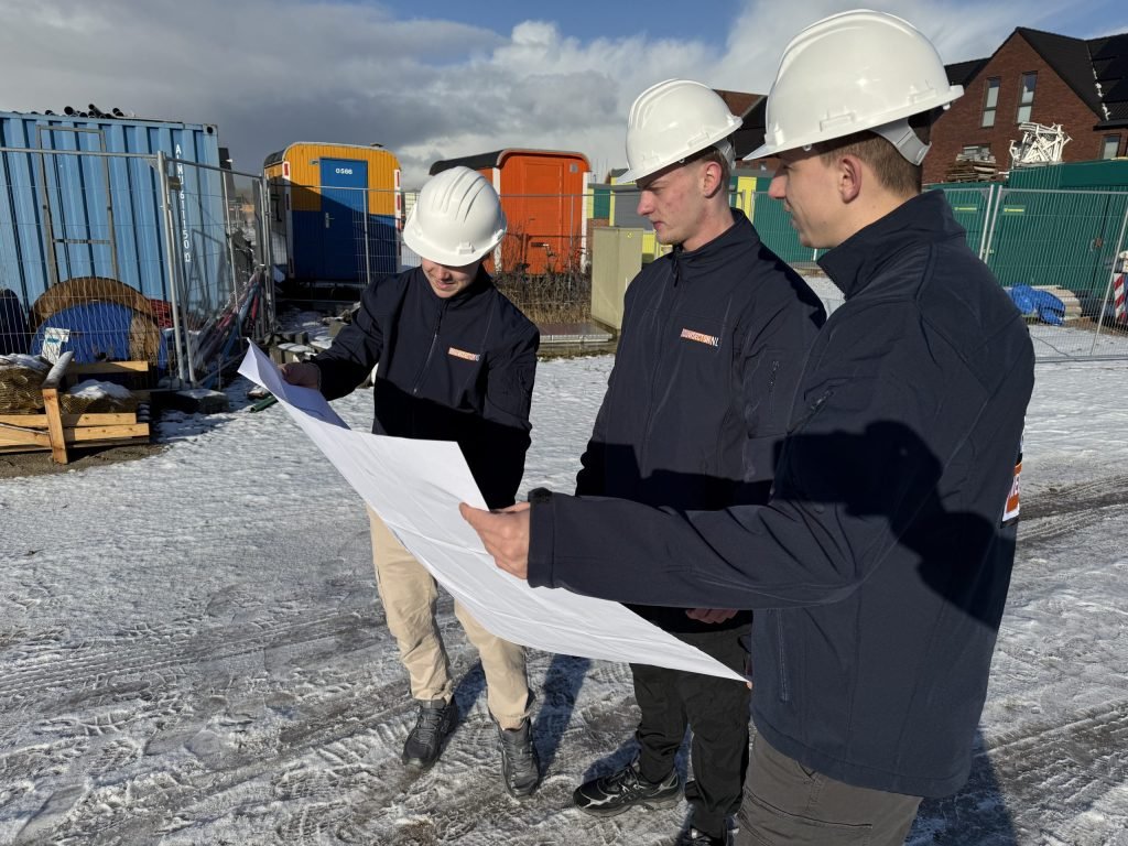 Drie vakmensen van Bouwsector Nederland met witte helm bekijken een tekening op een besneeuwde bouwplaats